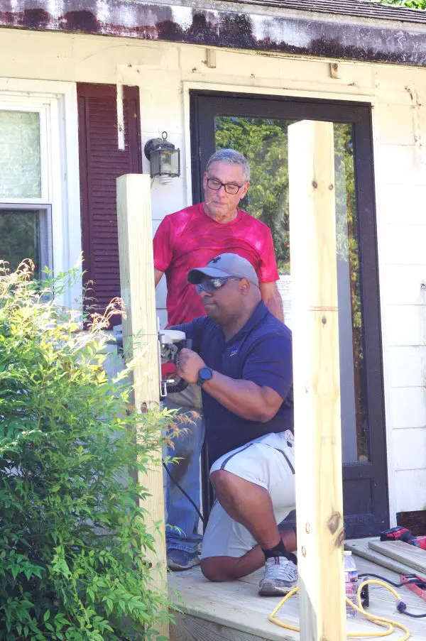 Two men outside a house, one standing and one crouching.