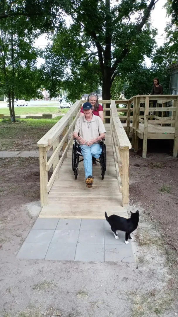 Elderly man sitting on a wooden ramp with a cat nearby.