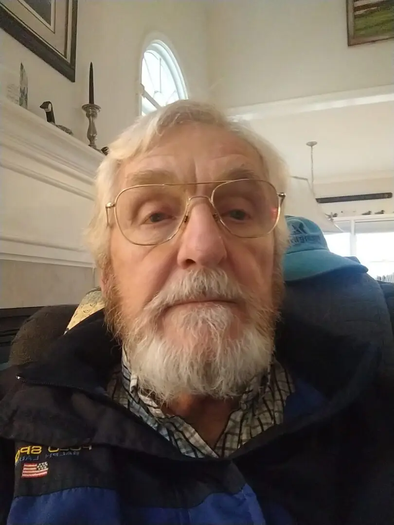 Elderly man with glasses and a beard sitting indoors.